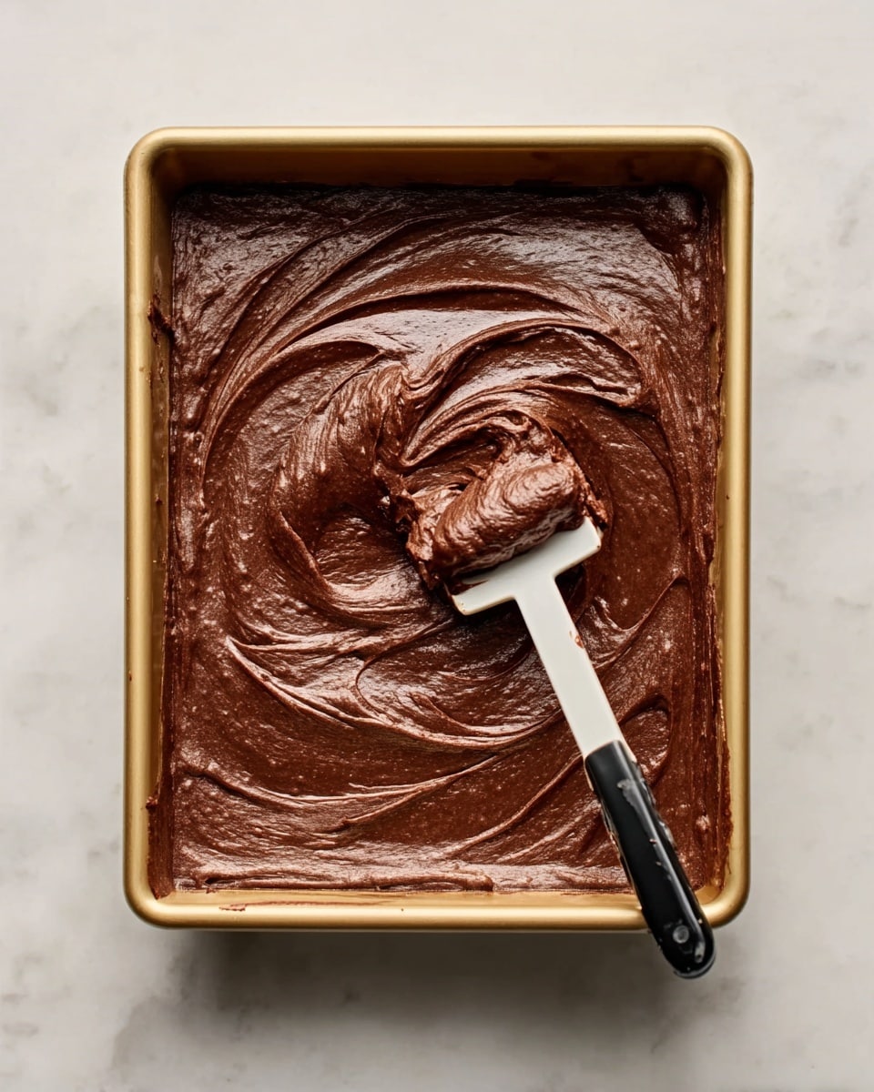 The image shows a rectangular golden baking pan filled with smooth, rich dark brown brownie batter. A white spoon with a black handle is scooping a portion from the center, creating swirled texture on the surface. The pan sits on a white marbled surface with soft lighting that highlights the glossy, thick consistency of the batter. Photo taken with an iphone --ar 4:5 --v 7