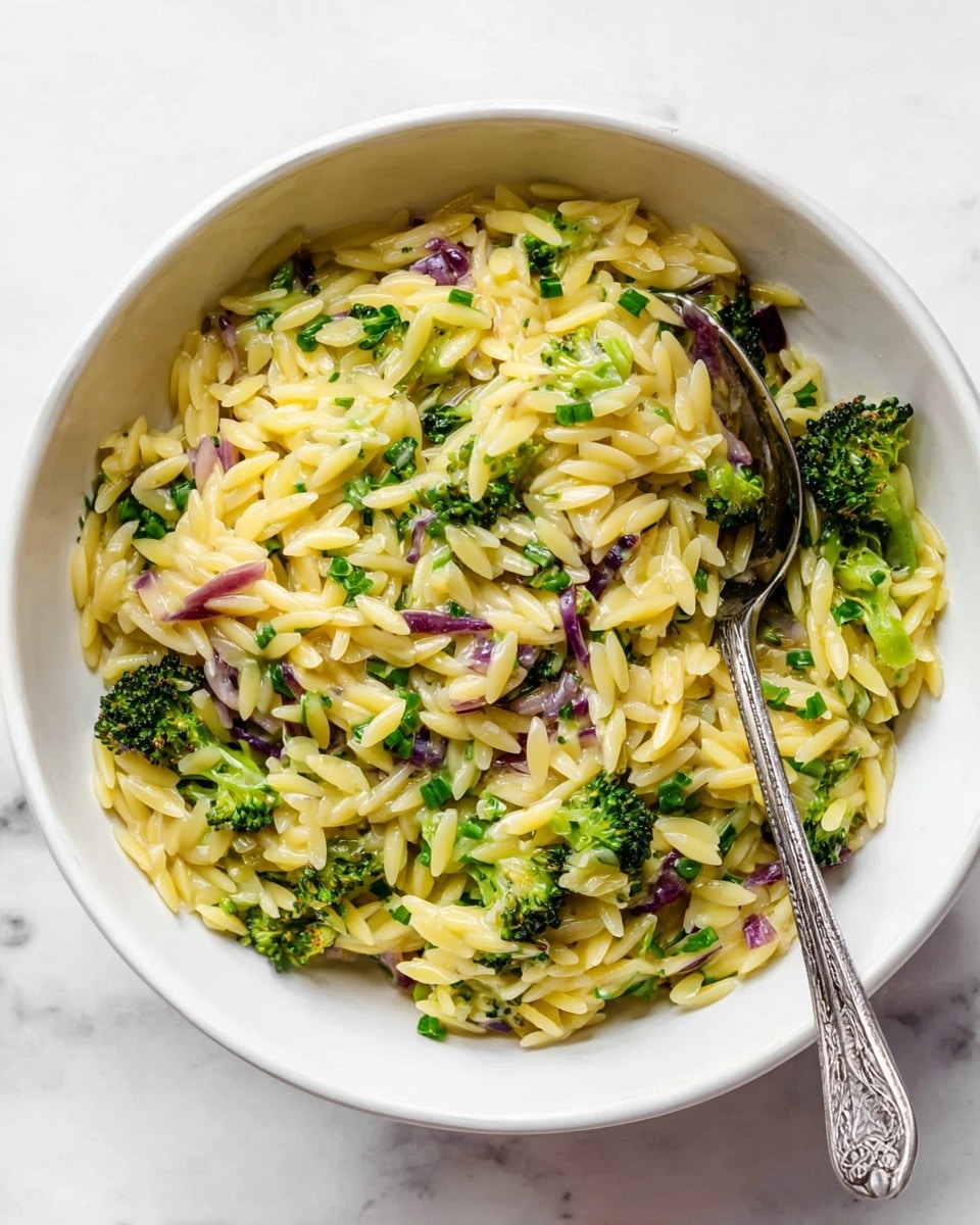 A white bowl with a mixture of yellow orzo pasta, small green broccoli pieces, and bits of purple onion. The orzo is creamy and shiny, filling most of the bowl, mixed evenly with the bright green broccoli florets and scattered purple onion chunks. A silver spoon rests inside the bowl, partly covered by the pasta. The bowl sits on a white marbled surface with soft natural light. photo taken with an iphone --ar 4:5 --v 7