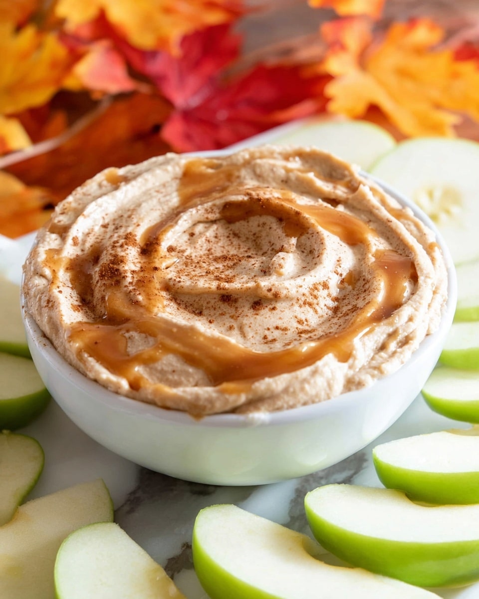 A white bowl filled with a thick, creamy dip that has a light brown color. The top of the dip has swirled ridges and is drizzled with a glossy caramel sauce, creating shiny, amber highlights. A sprinkle of cinnamon powder is spread unevenly over the caramel and dip surface. Surrounding the bowl are fresh green apple slices, light green with white inside, laying flat on a white marbled surface. In the background, blurred fall leaves in yellow, red, and orange tones add a cozy, autumn feel. Photo taken with an iphone --ar 4:5 --v 7