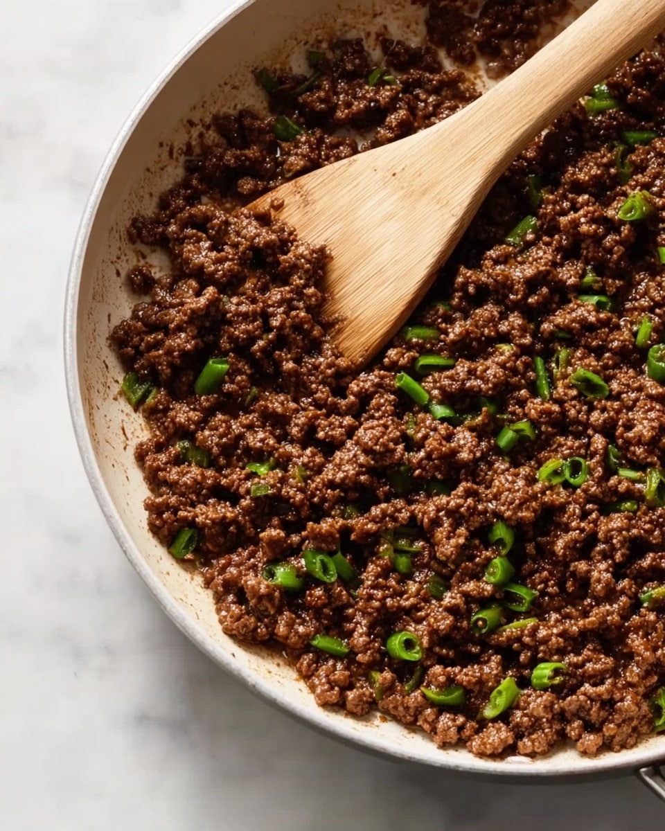 A close-up view of cooked minced meat browned in a white skillet with small green vegetable pieces mixed in, giving a fresh contrast to the deep brown meat. A wooden spoon with a light wood texture rests inside the pan, positioned diagonally with the handle on the left side. The skillet is on a white marbled surface that provides a clean, bright background. The meat looks juicy with a slight shine on the surface, and the green vegetable pieces are scattered evenly throughout. photo taken with an iphone --ar 4:5 --v 7