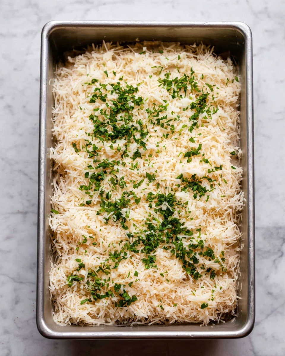 A rectangular metal baking pan holds a layered dish sitting on a white marbled surface. The bottom layer is light brown, topped with an even thicker layer of finely shredded, pale beige flakes, likely soft and fluffy in texture. Scattered evenly on top are small bits of bright green chopped herbs, adding color contrast and freshness to the pale layers beneath. The whole dish is contained within the pan and looks ready for baking. photo taken with an iphone --ar 4:5 --v 7
