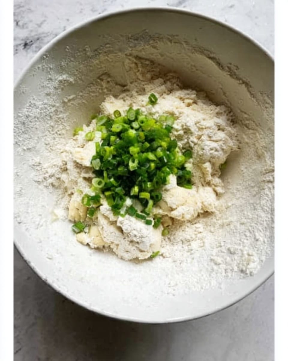 A close-up view of a white bowl with flour and small pieces of dough forming the bottom layer, topped with a small pile of chopped green onions in the center, all sitting on a white marbled surface. The textures show the flour dusted around the sides of the bowl with rough, uneven dough mixed in with the soft green onion pieces. Photo taken with an iphone --ar 4:5 --v 7