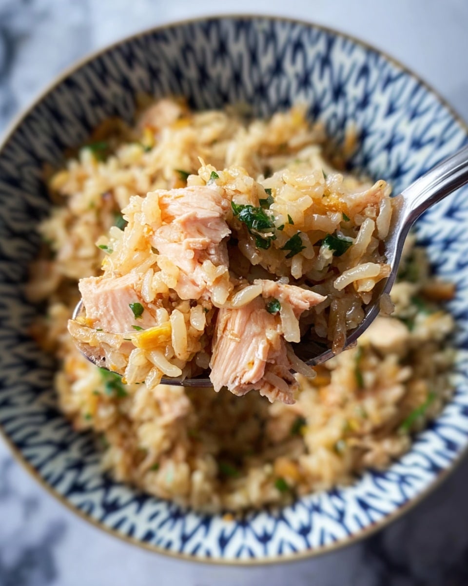 A close-up view of a spoon holding a mix of cooked rice and small pieces of light pink fish with some green herb bits scattered throughout, hovering above a white bowl with a blue geometric star-like pattern filled with more of the same rice and fish mixture. The rice is light brown with some small yellow pieces, and the fish looks tender and flaky, while some green herb pieces are sprinkled on top. The background surface is white marble. photo taken with an iphone --ar 4:5 --v 7