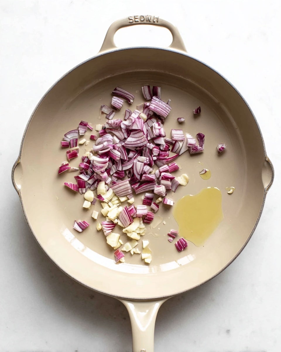 A beige pan with a single handle is shown from above on a white marbled surface, containing two layers of ingredients: the first layer is a small amount of light yellow oil spread slightly to the right, and the second layer is a mix of chopped red onions and small pieces of garlic placed mainly on the left side of the pan, the onions showing white and purple stripes with a firm texture and the garlic having a pale creamy white color, all ingredients sitting loosely together inside the pan photo taken with an iphone --ar 4:5 --v 7