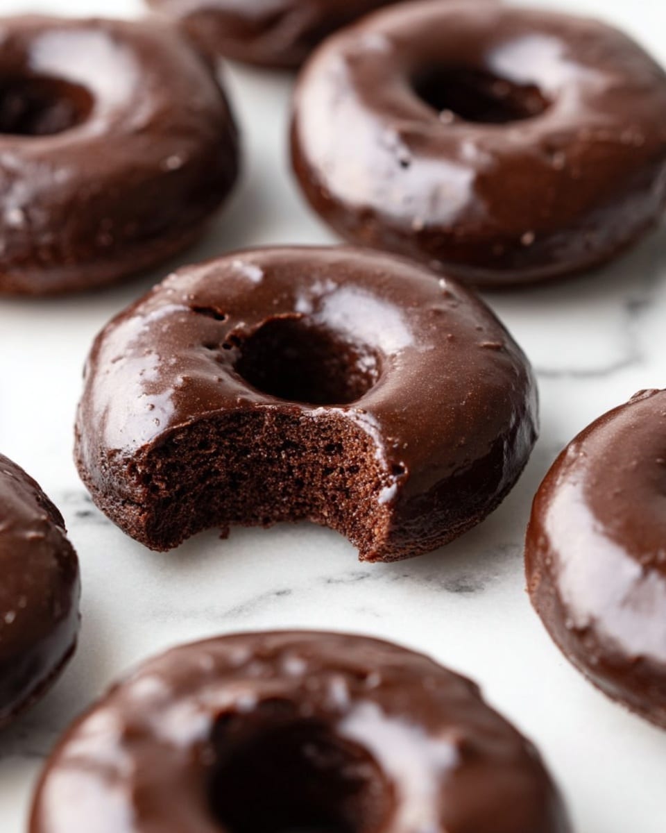 Several chocolate donuts with a dark brown, smooth glaze sit on a white marbled surface. One donut in the center has a bite taken out, showing a soft, moist, and dense interior that is a rich chocolate color. The donuts are round with a hole in the middle, and the glaze shines slightly, showing a delicate texture with small cracks. The focus is sharp on the bitten donut and softer on the surrounding donuts. Photo taken with an iphone --ar 4:5 --v 7