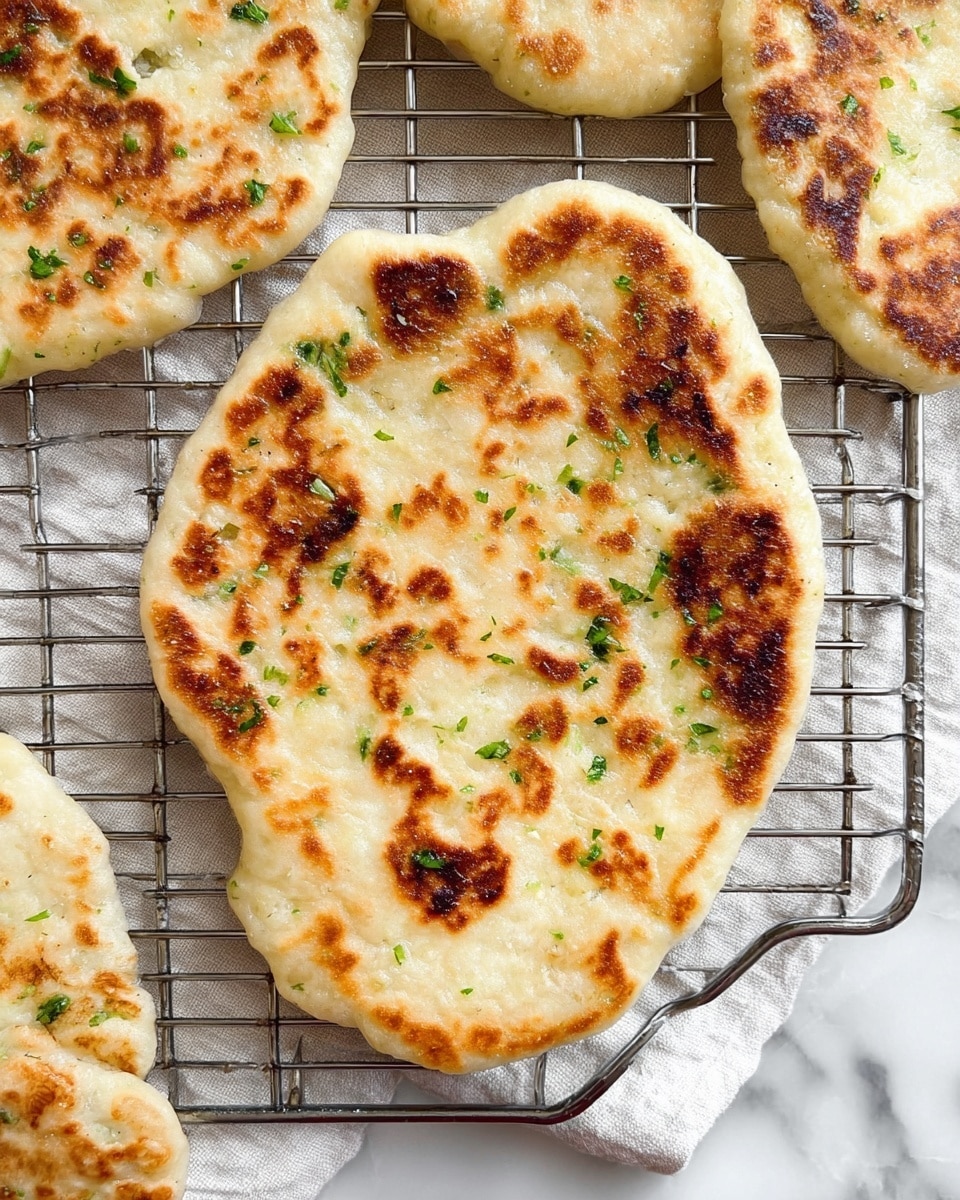 The image shows a piece of flatbread sitting on a metal rack above a white cloth. The flatbread has an uneven oval shape with a golden brown, toasted top that shows patches of light cream dough underneath. Small green bits, likely herbs, are scattered across the surface. The bread appears soft and fluffy with a slightly crispy texture from the toasting, and other similar flatbreads can be seen partially around the edges on the metal rack. The background has a white marbled texture. Photo taken with an iphone --ar 4:5 --v 7