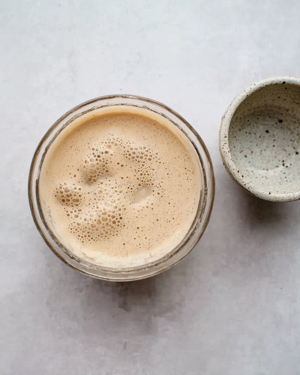 The image shows a clear glass bowl filled with a light beige, foamy liquid with bubbles on the surface. The liquid appears creamy and smooth with small uneven patches of foam scattered across. Next to the bowl, on a white marbled surface, is a small ceramic bowl or cup with a light beige and gray speckled pattern. The scene is simple and clean with soft lighting. Photo taken with an iphone --ar 4:5 --v 7