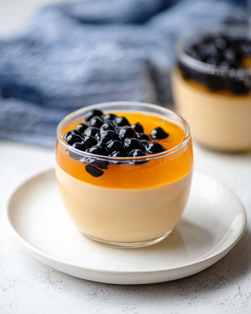 The image shows a clear glass cup sitting on a plain white plate, placed on a white marbled surface. The cup has three layers: the bottom layer is a smooth light beige creamy pudding filling most of the glass, the middle layer is a thin, bright orange jelly that covers part of the pudding, and the top layer is a cluster of shiny, round black tapioca pearls arranged on one side of the orange jelly. In the background, there is a blurred second glass with similar contents and a blue and white cloth. Photo taken with an iphone --ar 4:5 --v 7