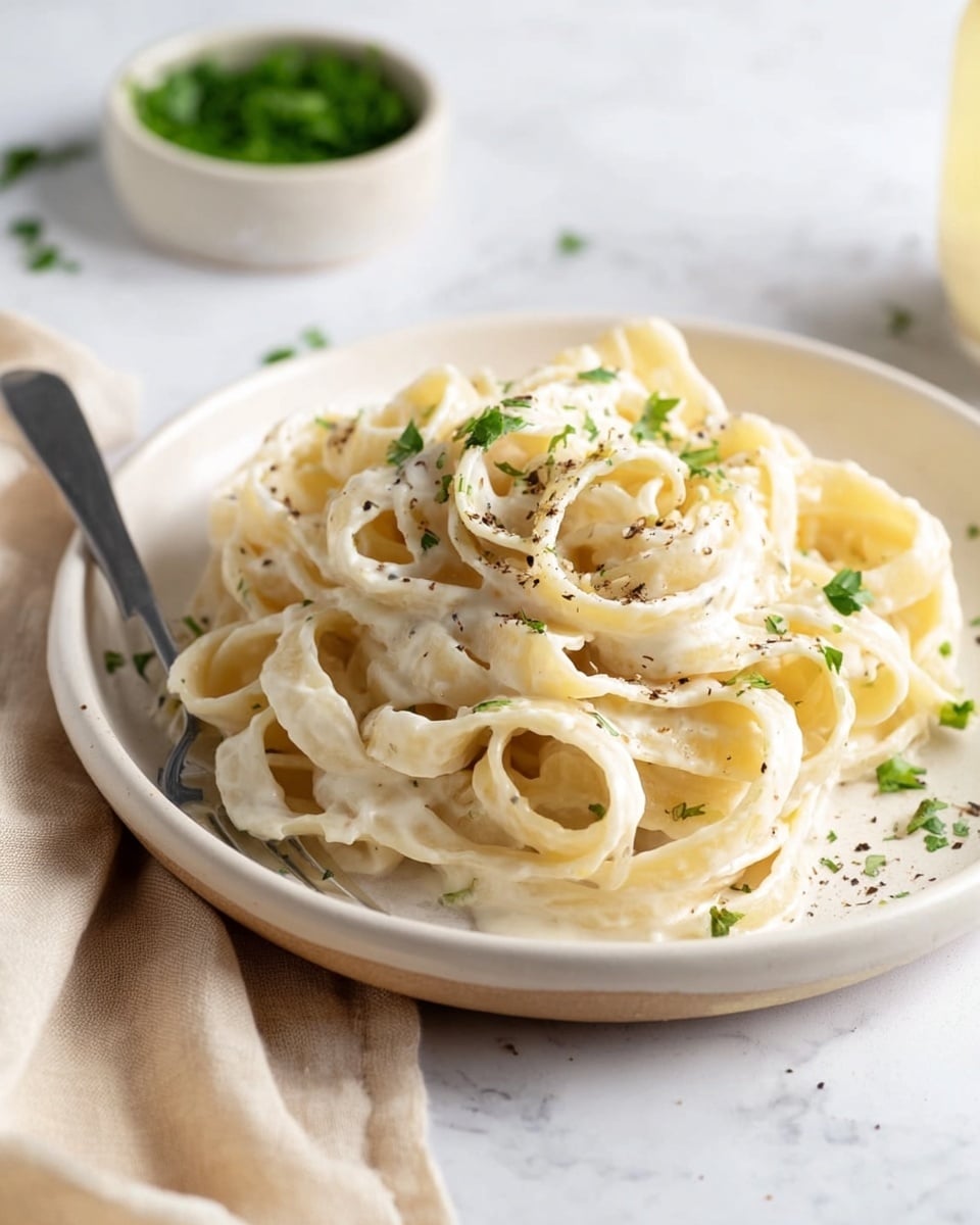 A white round plate holds a nest of creamy fettuccine pasta with a smooth white sauce, sprinkled evenly with small green herb bits and light black pepper flakes. The pasta strands are thick, soft, and curled, piled in a slightly messy way in the center. In the blurry background, there is a small white bowl filled with chopped green herbs on a white marbled surface. A fork is placed on the left edge of the plate, and a beige cloth with soft folds is in the lower corner. The overall look is fresh and simple, with soft natural light highlighting the food’s texture. photo taken with an iphone --ar 4:5 --v 7