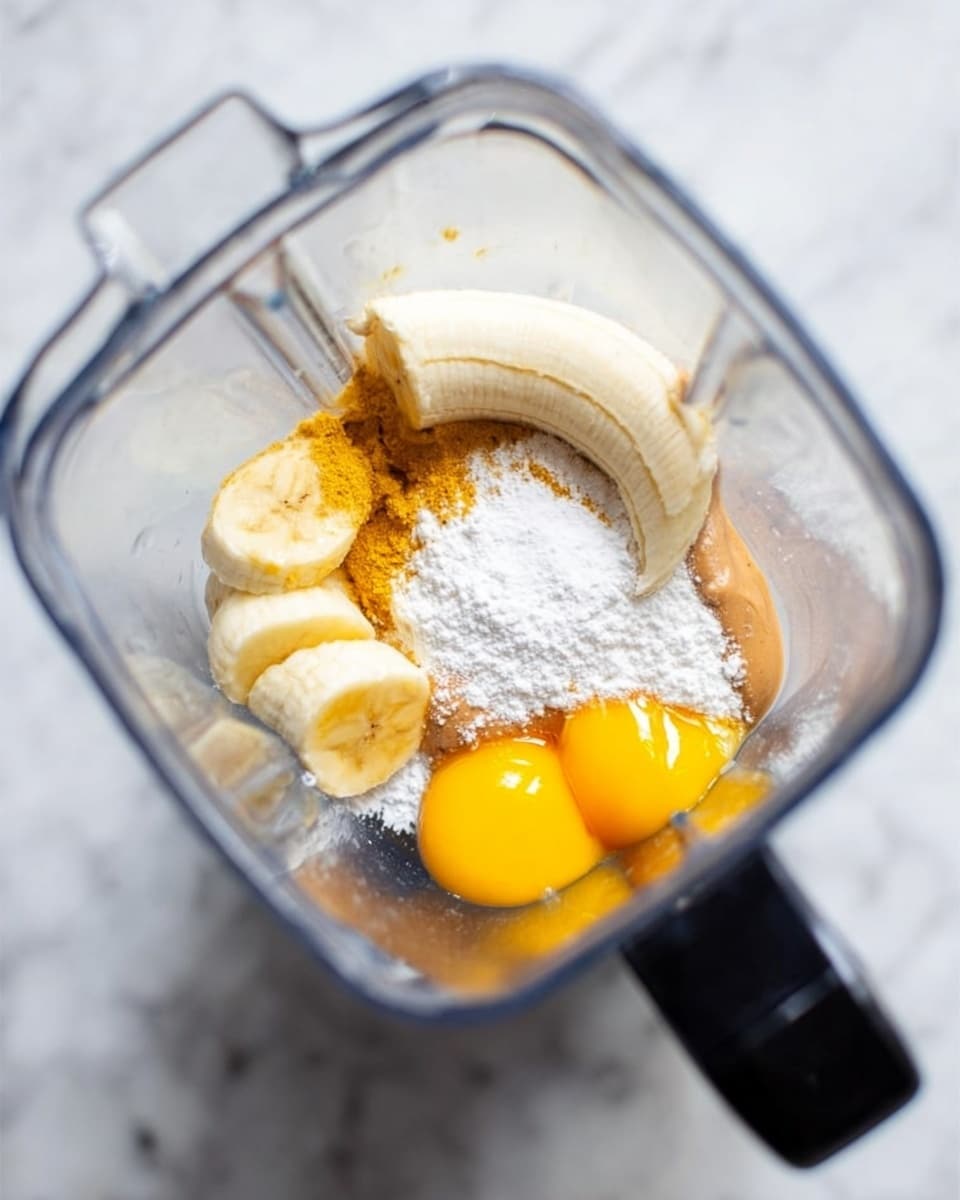 A clear blender jar sits on a white marbled surface. Inside, there are several layers of ingredients: a half peeled banana resting on one side, bright yellow egg yolks on the top right, a mound of white powder (likely flour or sugar) sprinkled in the center and toward the top, a light brown substance (possibly peanut butter) below the yolks, and some white powder around the brown layer. The edges of the jar are transparent, and the black handle is visible on the right side. The photo taken with an iphone --ar 4:5 --v 7