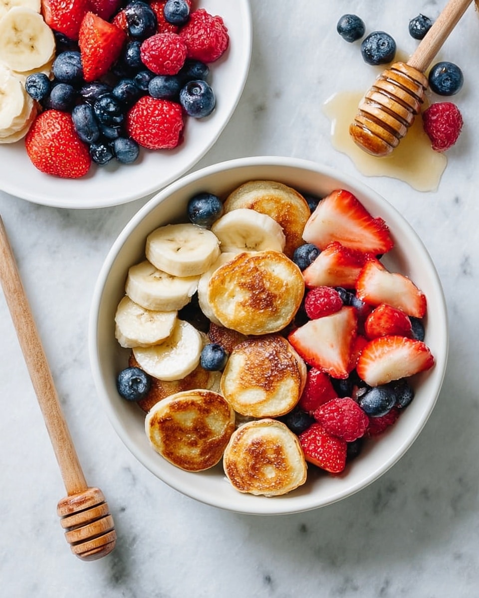 The image shows a white bowl filled with several small, round, golden-brown pancakes that have a slightly crispy texture. These mini pancakes form the base layer on one side of the bowl. On the other side, there is a colorful mix of fresh fruit slices including bright red strawberries, pale yellow banana slices, deep blue blueberries, and vibrant red raspberries, creating a fresh and inviting contrast. Next to the bowl, there is a white plate with more blueberries and strawberry pieces. A wooden honey dipper with honey drips is placed nearby on a white marbled textured surface, adding a natural touch to the scene. photo taken with an iphone --ar 4:5 --v 7
