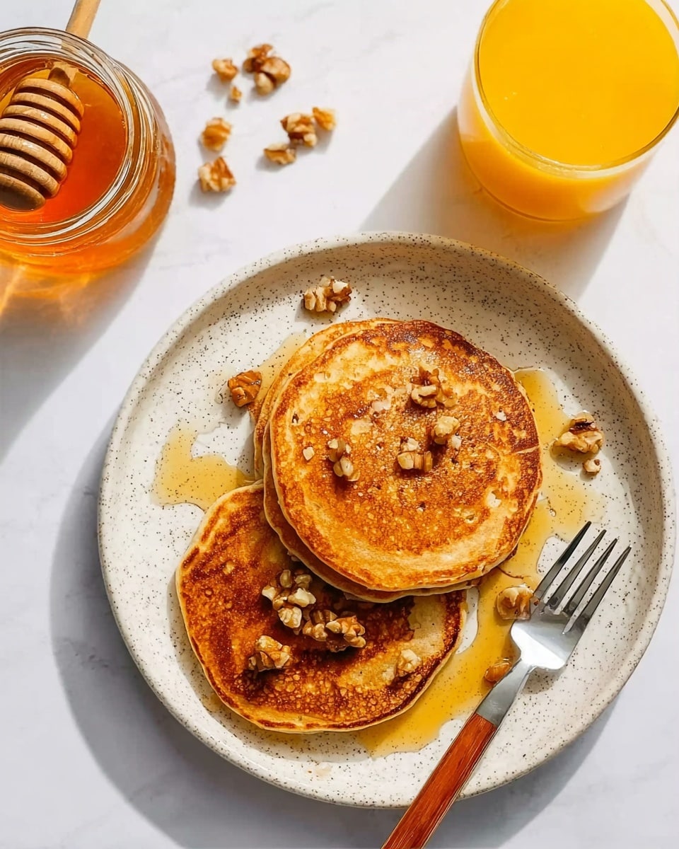 A white speckled plate holds three golden brown pancakes stacked on top of each other, topped with small pieces of chopped walnuts and drizzled with honey that shines under the light. On the right side of the plate lies a fork with a brown wooden handle. To the top left next to the plate, there is a clear jar filled with honey with a honey dipper resting inside it. At the bottom left of the image is a glass full of bright orange juice. The background is a smooth white marbled surface. Photo taken with an iphone --ar 4:5 --v 7
