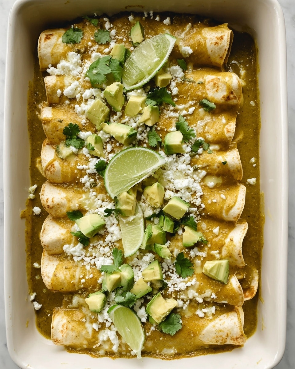 This image shows a white baking dish filled with two rows of eight rolled tortillas, covered with a golden-green sauce that has a smooth, slightly thick texture. On top of the tortillas, there are scattered layers of white crumbled cheese, small bright green avocado cubes, and fresh cilantro leaves, adding contrast and freshness. Lime wedges, cut into quarters, are placed on the top layer, adding bright pops of green color. The dish sits on a white marbled surface that enhances the colors of the food. photo taken with an iphone --ar 4:5 --v 7