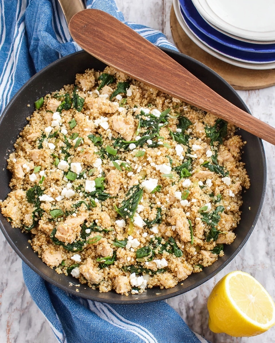 A black skillet filled with cooked quinoa mixed with small pieces of light beige chicken, bright green spinach leaves, and white crumbles of cheese. The mix has a slightly grainy texture from the quinoa, scattered evenly with the chicken and spinach. On top, a wooden spoon rests diagonally, showing its brown wood grain. Nearby, a white marbled surface holds a folded blue cloth with white stripes and a white plate stacked with blue plates. A lemon half with a bright yellow color sits at the side, adding a fresh touch to the scene. photo taken with an iphone --ar 4:5 --v 7