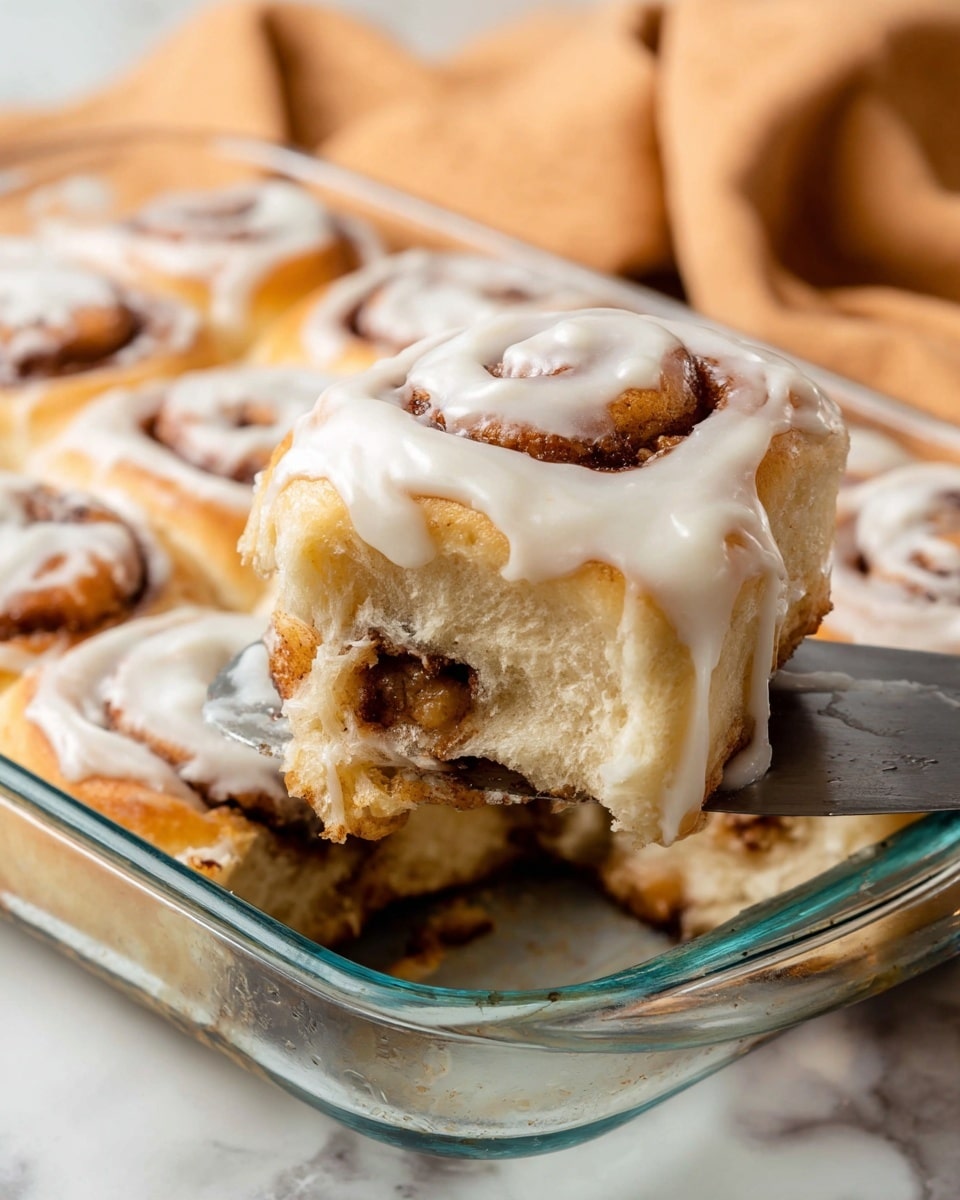 The image shows a close-up of a freshly baked cinnamon roll being lifted with a metal spatula from a clear glass dish filled with more cinnamon rolls. The cinnamon roll has three visible layers: the outer layer is a golden-brown dough with a soft, fluffy texture, the middle layer has a swirl of dark brown cinnamon filling, and the top layer is a thick, creamy white icing generously spread over the roll, dripping slightly on the sides. The background has a soft white marbled texture with a blurred light brown cloth. Photo taken with an iphone --ar 4:5 --v 7