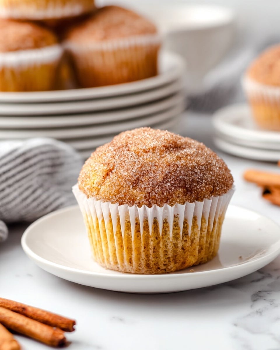 A single cinnamon sugar muffin sits in a white round dish, showing a light golden brown cake base with a crinkled white paper liner wrapping it. The muffin top is rounded and textured with a sparkling layer of cinnamon sugar, giving it a rough, grainy look. In the background, more muffins rest on stacked white plates and bowls on a white marbled surface, with a striped gray and white cloth and a few cinnamon sticks beside them. The warm light adds softness and highlights the sugar crystals on top. photo taken with an iphone --ar 4:5 --v 7