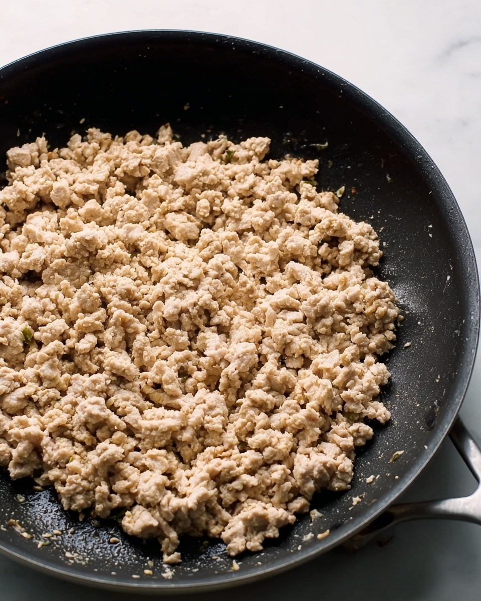 A close-up view of cooked ground meat with a light beige color and slightly crumbly texture spread evenly in a large black pan. The pan has a matte finish with subtle sheen from cooking oils, showing some moisture glistening on the meat. The edges of the pan are rounded, with a handle visible on the left side. The background is a white marbled surface. photo taken with an iphone --ar 4:5 --v 7