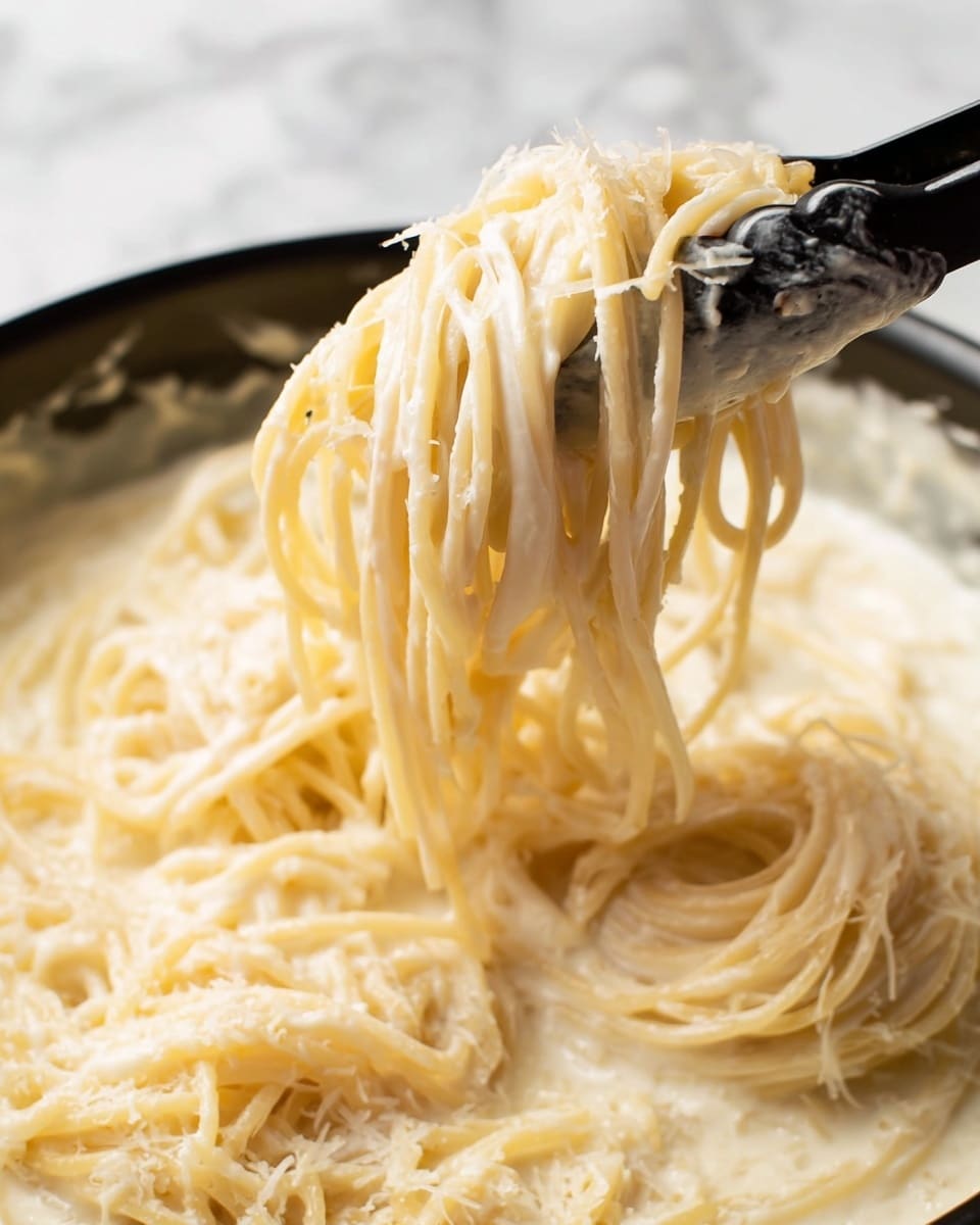 The image shows a close-up of creamy spaghetti pasta being lifted by black tongs in a pan. The spaghetti strands are soaked evenly in a smooth, white sauce that looks thick and rich. The pasta appears soft and well-coated with the sauce, with some grated cheese sprinkled on top, adding a slightly grainy texture. The background is a white marbled texture, and the focus is tight on the pasta and sauce, showing the shine and creaminess clearly. Photo taken with an iphone --ar 4:5 --v 7