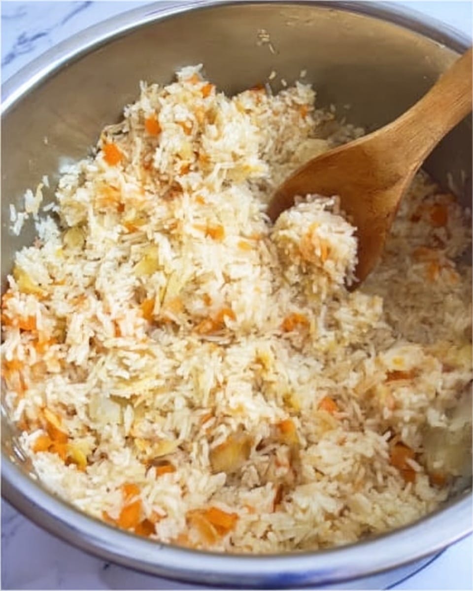 A close-up view inside a large silver pot showing a mixed rice dish with visible orange vegetable pieces and small bits of white and light brown ingredients, stirred with a wooden spoon. The texture looks soft and slightly fluffy, with the ingredients evenly spread throughout the rice. The background surface is a white marbled texture. photo taken with an iphone --ar 4:5 --v 7