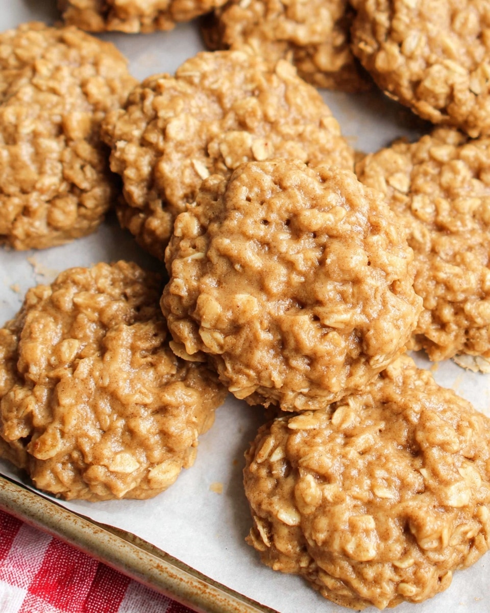 A close-up view of several oatmeal cookies on a white parchment paper lined baking tray, showing their light golden brown color. Each cookie has a rough and chunky texture with visible oat flakes throughout. The cookies are round and slightly uneven in shape, overlapping one another. The baking tray edges are visible in the bottom left and right corners, and the background surface is a white marbled texture with a hint of a red and white checkered cloth. Photo taken with an iphone --ar 4:5 --v 7