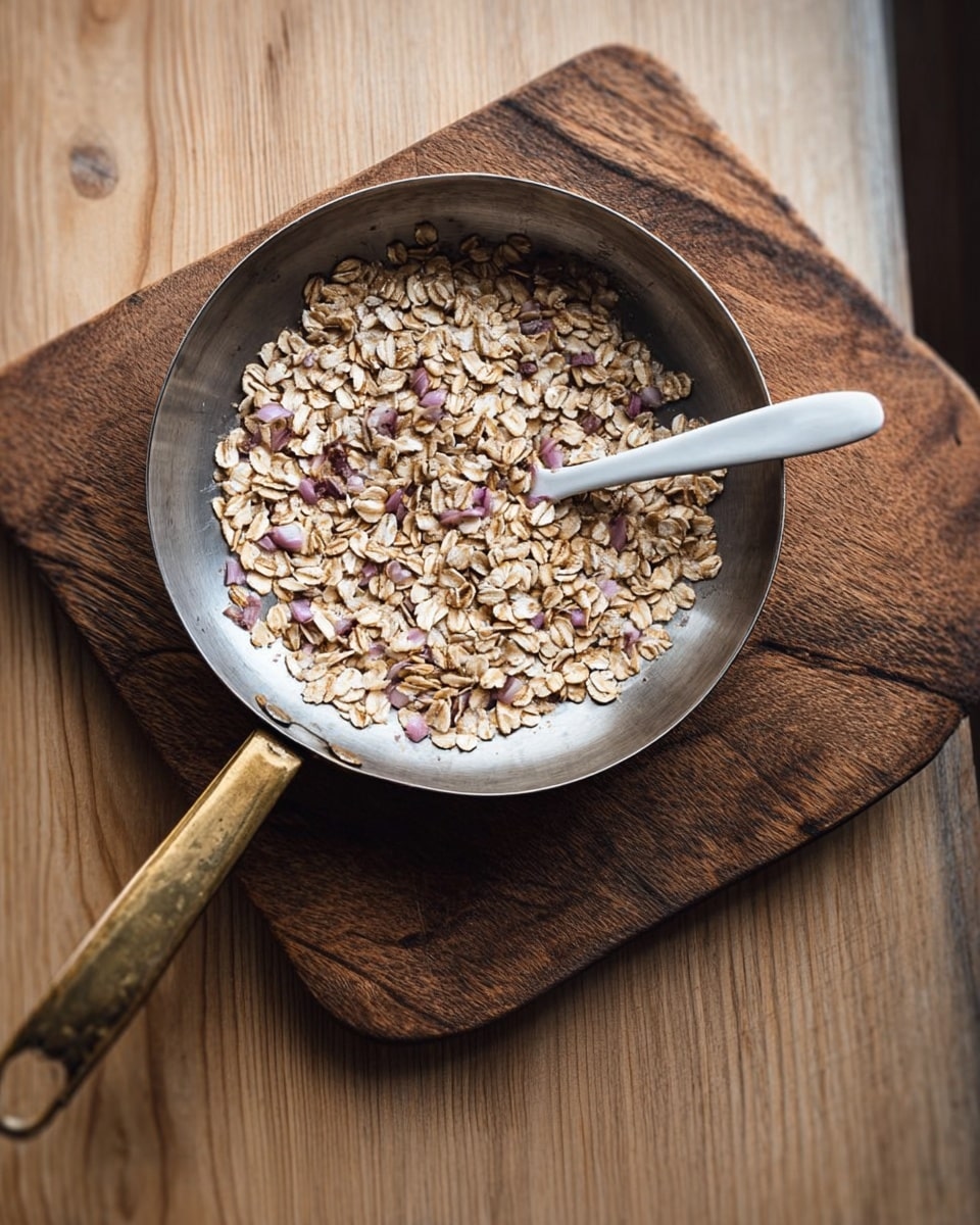 A round metal pan with a shiny, slightly worn surface holds a single layer of light brown oats mixed with small pieces of pale purple onion. A white ceramic spoon rests on top of the oats near the center of the pan. The pan has a long brass handle pointing diagonally down to the right. The pan sits on a wooden board with a rich brown grain, which is placed on a light wood table. The overall setting shows natural soft light highlighting the textures. photo taken with an iphone --ar 4:5 --v 7