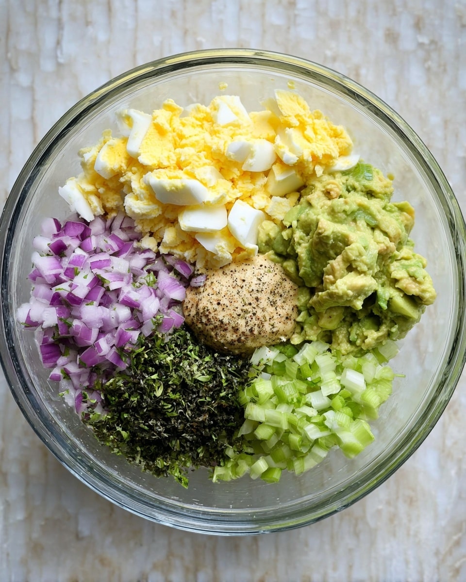 A clear glass bowl holds a mix of six colorful layers arranged side by side: bright yellow and white chopped boiled eggs at the bottom left, finely diced purple onions with green herb sprinkles just above it, a mound of dark green dried herbs near the top, creamy light green mashed avocado on the right, pale green chopped celery below the avocado, and a small dollop of light brown mustard with black pepper speckles in the center. The bowl sits on a white marbled textured surface photo taken with an iphone --ar 4:5 --v 7