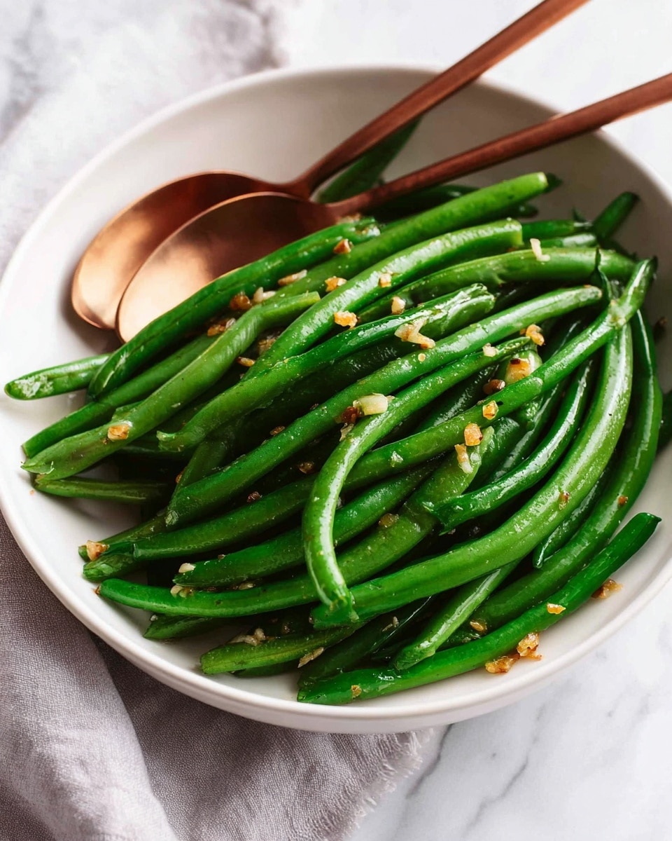 A white round shallow bowl filled with bright green steamed green beans, each bean smooth and slightly shiny with a fresh texture. Small bits of golden brown garlic scattered on top, adding warm color contrast. Two copper utensils, a fork and a spoon, rest inside the bowl leaning against the beans. The bowl sits on a white marbled surface with a light gray cloth partially visible on the left side. photo taken with an iphone --ar 4:5 --v 7