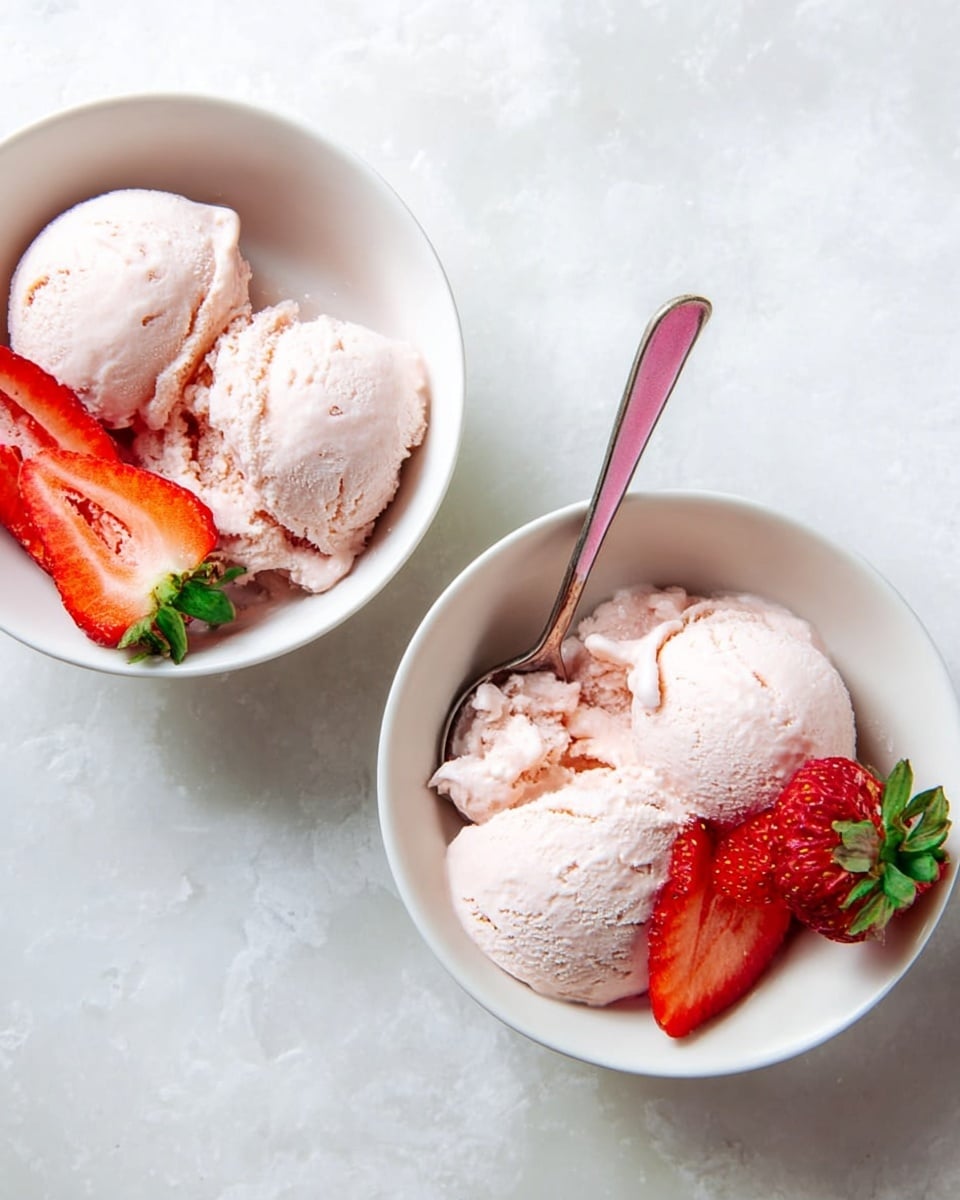 Two white bowls sit on a white marbled surface. Each bowl has two scoops of light pink creamy ice cream with a smooth texture. One bowl has a spoon with a pink handle inside, scooping some ice cream. Both bowls have fresh red strawberry halves with green leaves placed next to the ice cream, adding a bright color contrast. The photo looks natural and bright, photo taken with an iphone --ar 4:5 --v 7