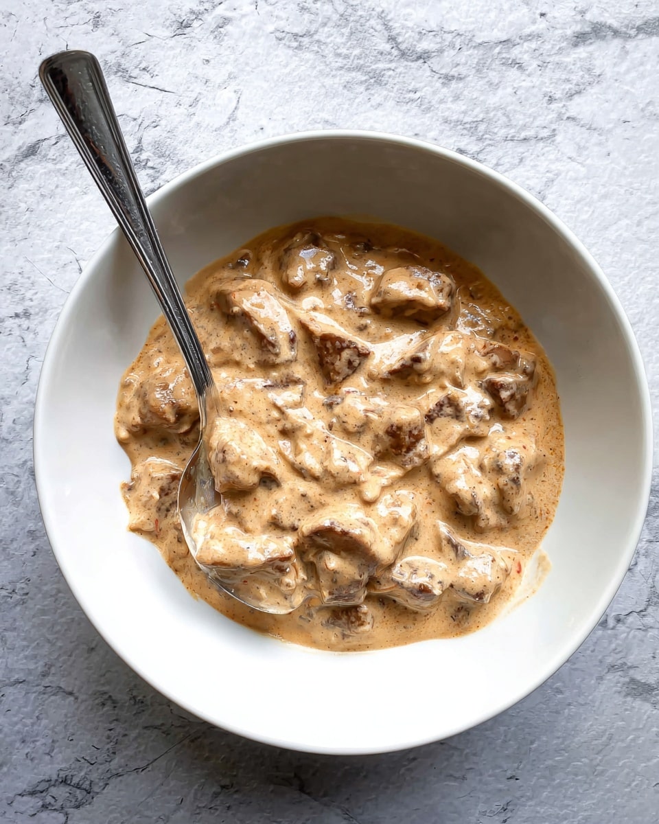 A white bowl filled with thick, creamy sauce that has a light brown color with darker brown specks, covering irregular chunk pieces of meat beneath it. A silver spoon is placed inside the bowl on the left side, partially submerged in the sauce. The bowl sits on a surface with a white marbled texture. photo taken with an iphone --ar 4:5 --v 7