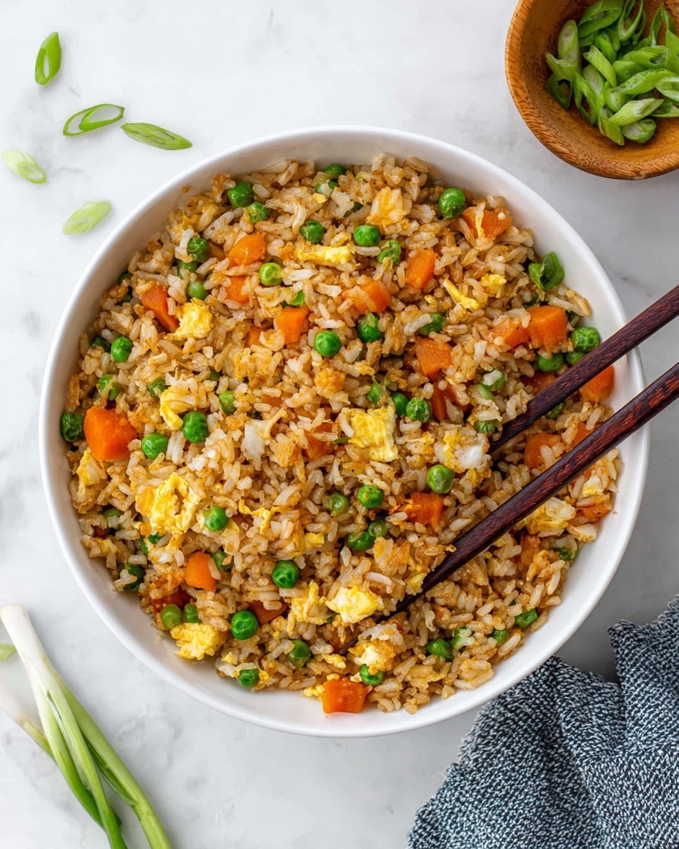 A white bowl filled with one layer of rice mixed with small green peas, orange carrot cubes, and yellow scrambled egg pieces, all evenly mixed and slightly shiny from light soy sauce. The rice is brownish with a mix of textures from soft rice grains to firmer vegetables and eggs. Two dark wooden chopsticks rest on the right side of the bowl, partly inside the rice. Around the bowl, some green peas and slices of green onion are scattered on a white marbled surface. A small wooden bowl with more sliced green onions is at the top right corner, and a blue-gray textured cloth lies at the bottom left corner. Photo taken with an iphone --ar 4:5 --v 7