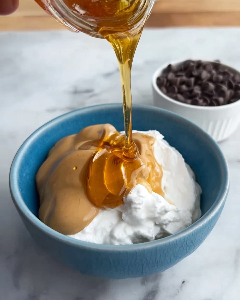A close-up of a blue bowl filled with three visible layers: white cottage cheese on the right, a smooth light brown peanut butter layer in the center, and golden honey being poured on top forming shiny drips down the sides. The bowl sits on a white marbled surface with a small white bowl of dark chocolate chips slightly blurred in the top right corner. A woman's hand is holding the container pouring the honey. photo taken with an iphone --ar 4:5 --v 7