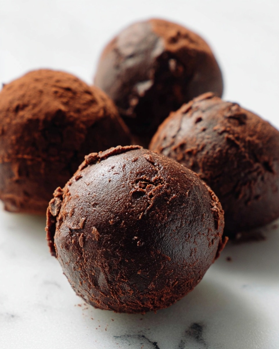 The image shows four dark brown balls of chocolate dough or truffle, each with a rough and crumbly texture. The balls are placed close together on a white marbled surface, with one ball in the front clearly in focus and the other three slightly blurred at the back. The chocolate balls have an uneven surface with small cracks and some glossy spots reflecting light. Photo taken with an iphone --ar 4:5 --v 7