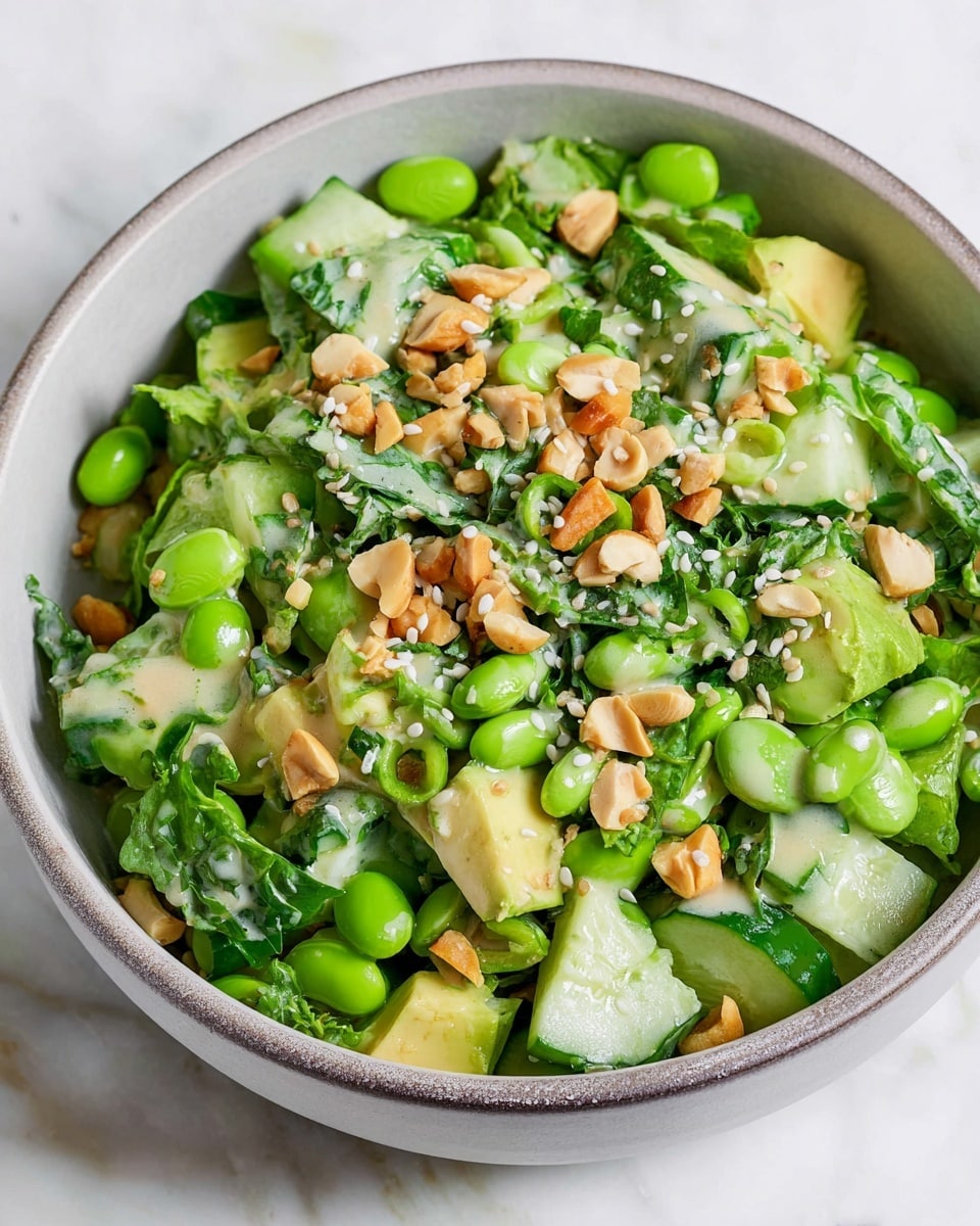 A close-up image of a fresh salad in a white bowl placed on a white marbled surface. The salad has many layers with bright green edamame beans as the base, mixed with chopped cucumber pieces that show a green outer skin and a lighter green inside. There are small chunks of avocado giving a creamy yellow-green color, along with chopped dark green leafy herbs evenly spread throughout. On top, there are light brown nuts scattered for texture, and a light creamy dressing drizzled over the salad with white sesame seeds sprinkled across the whole dish. The bowl has a grayish tone inside with a simple rim, and the entire setting looks fresh and clean. photo taken with an iphone --ar 4:5 --v 7