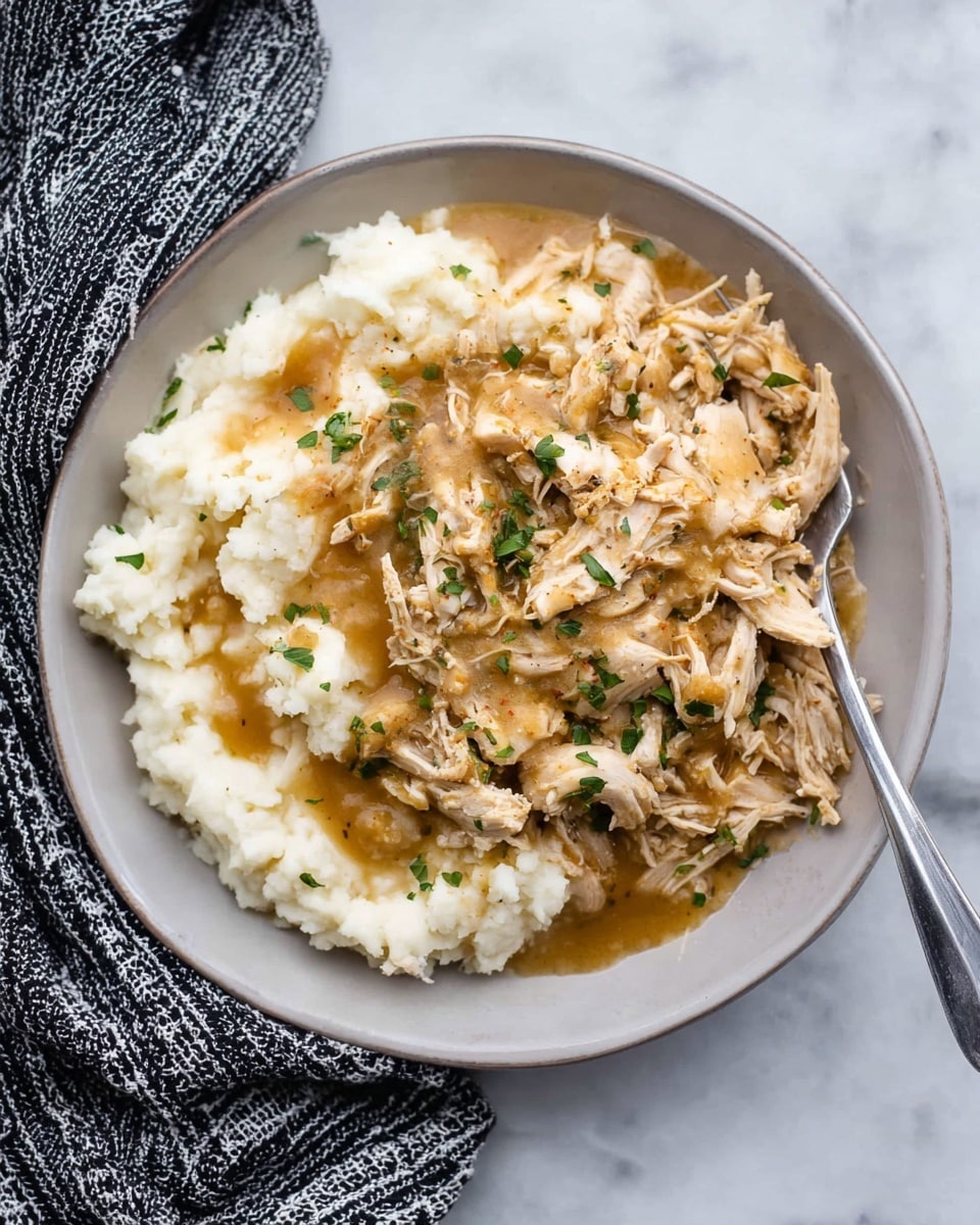 A white plate holds two main layers: the bottom layer is creamy mashed potatoes with a smooth and slightly lumpy texture, spread evenly across half the plate, in a soft off-white color. On top, there is a generous layer of shredded chicken mixed with light brown gravy, giving it a moist, glossy look. Small bits of green herbs are sprinkled over the chicken and mashed potatoes. A silver fork rests on the plate’s right side, partially under the shredded chicken. The dish is set on a white marbled surface with a black-and-white patterned cloth placed near the plate. photo taken with an iphone --ar 4:5 --v 7