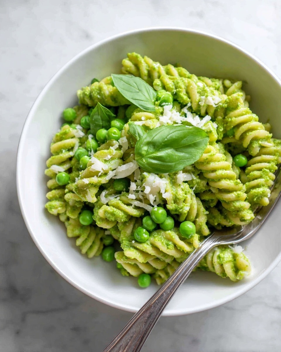 A white bowl filled with three layers of green pasta spirals coated in bright green pesto sauce mixed with fresh green peas scattered throughout; the pasta is topped lightly with small white cheese sprinkles and a few fresh green basil leaves as garnish. A silver fork rests on the right side inside the bowl, all placed on a white marbled surface. photo taken with an iphone --ar 4:5 --v 7