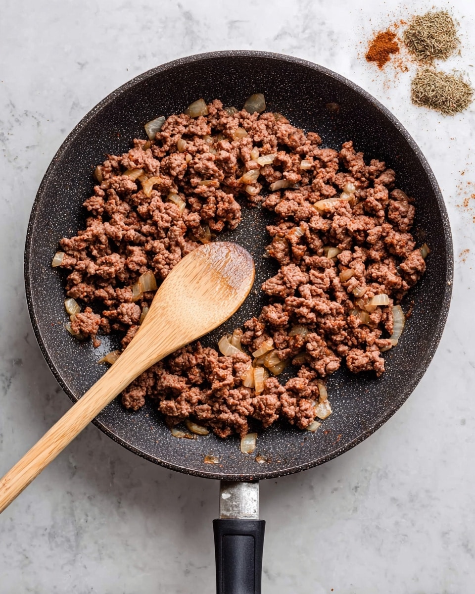 A top view of a black frying pan with a textured surface holding cooked ground beef mixed with diced onions; the beef is crumbly and brown with bits of translucent cooked onion spread evenly. A long wooden spoon with a smooth texture rests inside the pan, touching the meat and extending outward from the pan's right side. The pan has a black handle that lies flat on a white marbled surface. Small piles of brown spices can be seen in the top right corner of the surface photo taken with an iphone --ar 4:5 --v 7