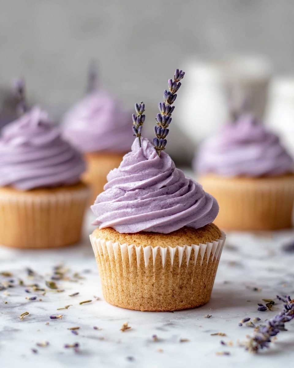 The image shows a close-up of a light brown cupcake with a smooth, purple swirl of frosting on top, decorated with two thin dried lavender flower stems sticking out of the frosting. In the background, there are four more similar cupcakes, each with the same purple frosting and lavender decoration, slightly blurred. The cupcakes are placed on a white marbled surface scattered with small dried lavender flowers. The lighting is soft and natural, highlighting the texture of the frosting and the cupcake base. photo taken with an iphone --ar 4:5 --v 7