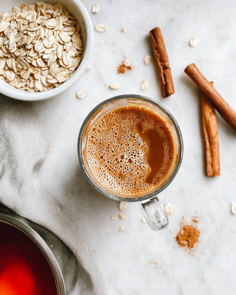 A clear glass cup sits in the center on a white marbled surface, filled with a warm brown drink that has a thick layer of bubbly foam on top, showing mixed lighter and darker brown colors with a creamy texture. To the top left is a white bowl filled with light tan rolled oats, some spilled oats scattered around nearby. Cinnamon sticks lay on the surface near the bowl, showing rich brown color and a rough texture. A stainless steel pot edge with reddish liquid and a floating cinnamon stick is partially visible on the left side. The cozy setup has a light grey cloth nearby adding a soft texture. photo taken with an iphone --ar 4:5 --v 7