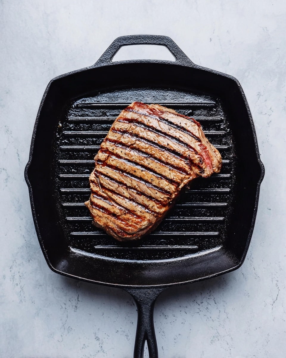 A single large grilled steak sits centered in a black cast iron grill pan, showing dark brown sear marks in parallel lines across its light brown surface with slightly darker edges. The pan has a broad handle at the top, and the whole scene rests on a white marbled surface that creates a clean, bright background. Photo taken with an iphone --ar 4:5 --v 7
