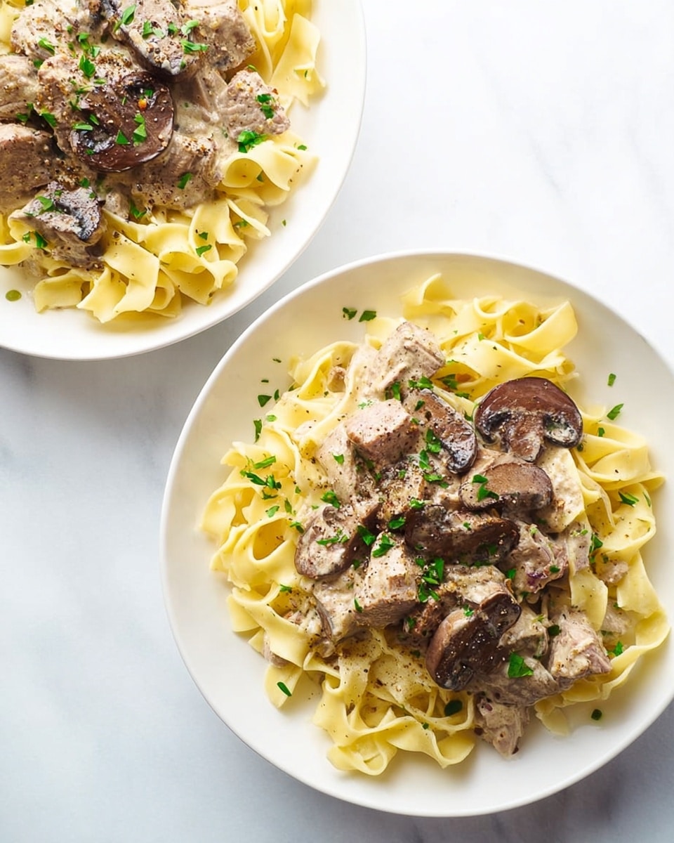 Two white plates sit on a white marbled surface, each filled with a bed of pale yellow wide egg noodles arranged loosely with soft folds. On top of the noodles is a creamy sauce with chunks of cooked meat and thick slices of brown mushrooms, seasoned lightly with black pepper. Small pieces of green herbs are scattered over the dish, adding a fresh touch of color. The creamy sauce looks rich but smooth, coating the meat and mushrooms without overwhelming the pasta. Photo taken with an iphone --ar 4:5 --v 7