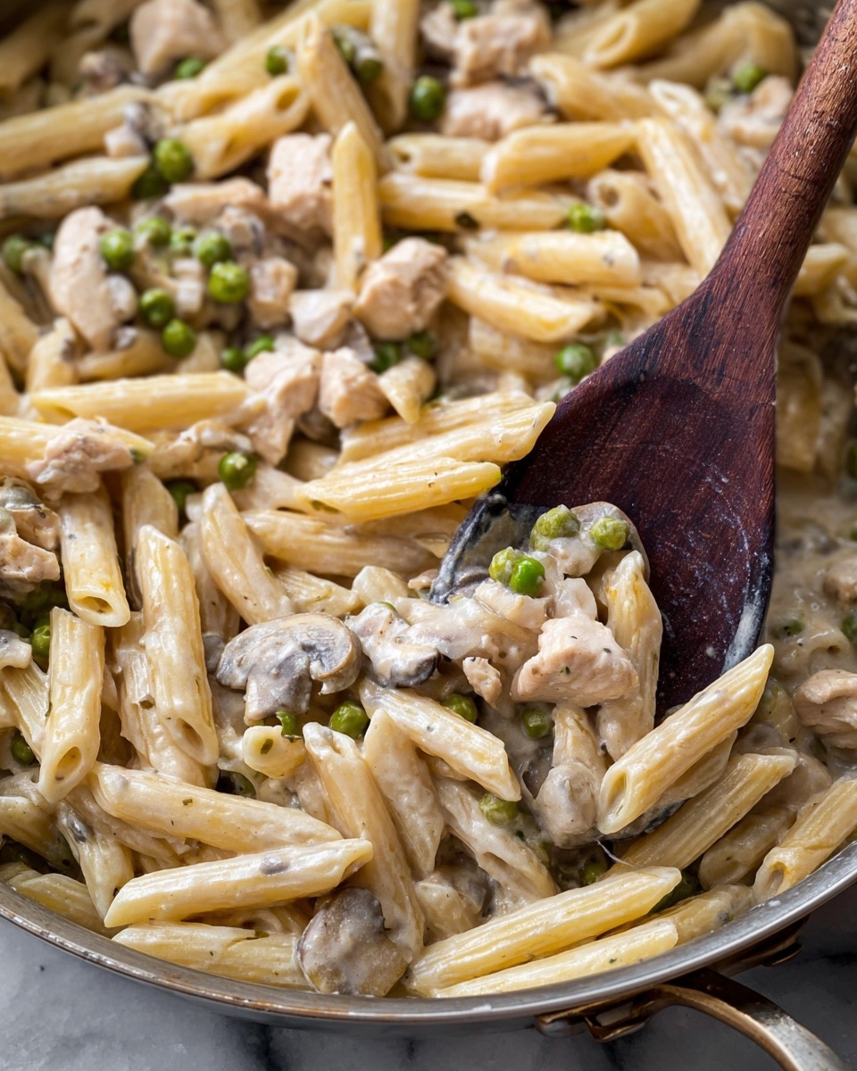 A close-up view of a creamy pasta dish in a metal pan showing one main layer of short, tube-shaped penne pasta colored light beige with a smooth, shiny cream sauce coating each piece. Scattered throughout the pasta are small pieces of light beige chicken and green peas, with some pieces of sliced mushrooms in muted brown and gray tones. A dark wooden spoon scoops part of the pasta, highlighting the glossy texture of the sauce and ingredients. The background is a white marbled texture. photo taken with an iphone --ar 4:5 --v 7