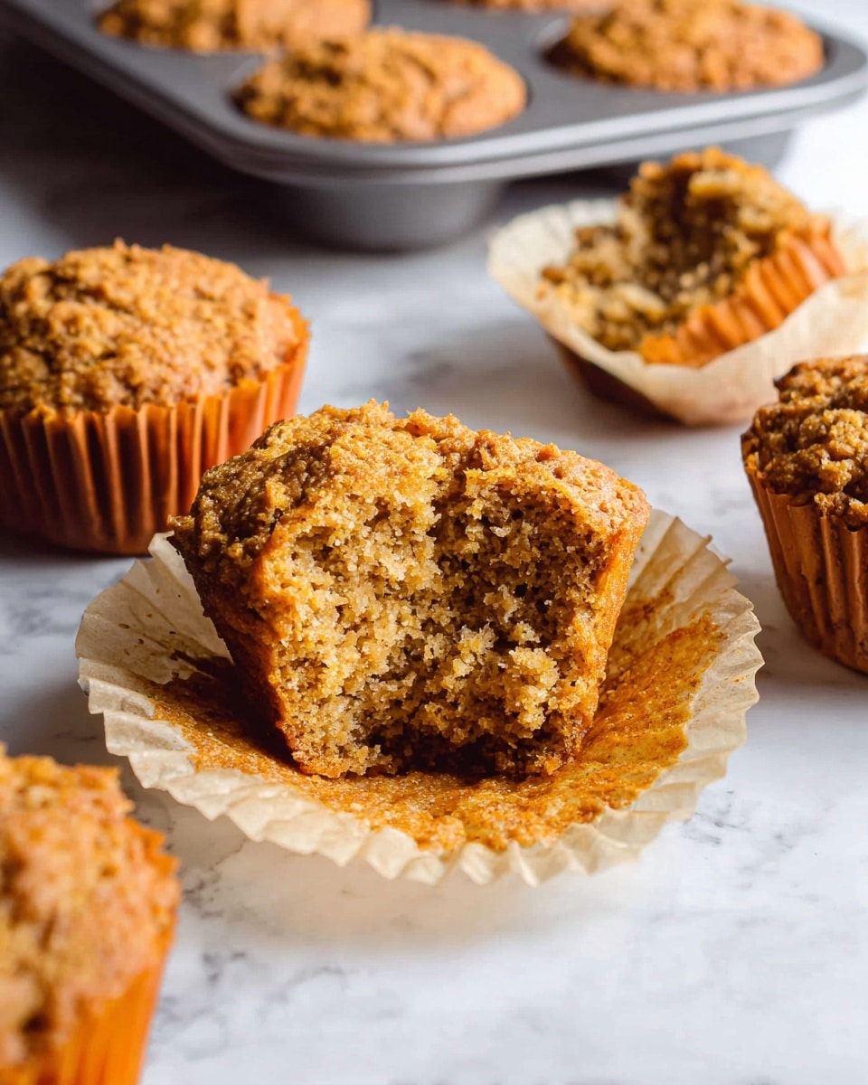 The image shows a close-up of several light brown muffins with a crumbly texture on a white marbled surface. One muffin is in the center, partially unwrapped from its beige paper liner, with a bite taken out of it revealing a dense, grainy inside. Other whole muffins in paper liners sit around the main muffin, and an orange-tinted muffin tray holds more muffins in the background. The light is soft, highlighting the rough surfaces and moist textures of the muffins. photo taken with an iphone --ar 4:5 --v 7