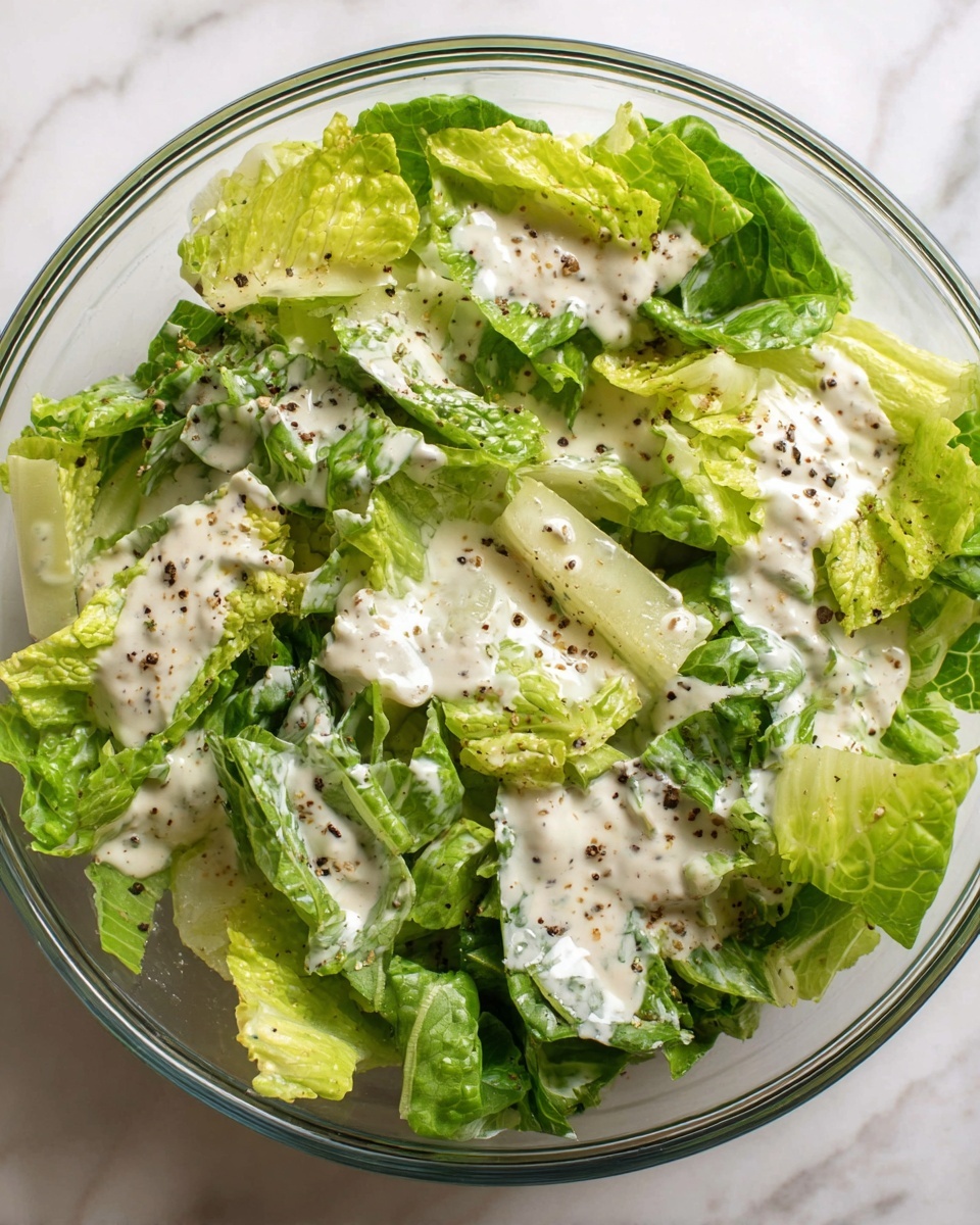A clear glass bowl filled with fresh green lettuce leaves as the base layer, showing a mix of light green to dark green shades with crisp textures. The lettuce is generously covered by a creamy white dressing that spreads unevenly over the leaves, with black pepper flakes sprinkled lightly on top for contrast. The bowl sits on a white marbled surface. photo taken with an iphone --ar 4:5 --v 7