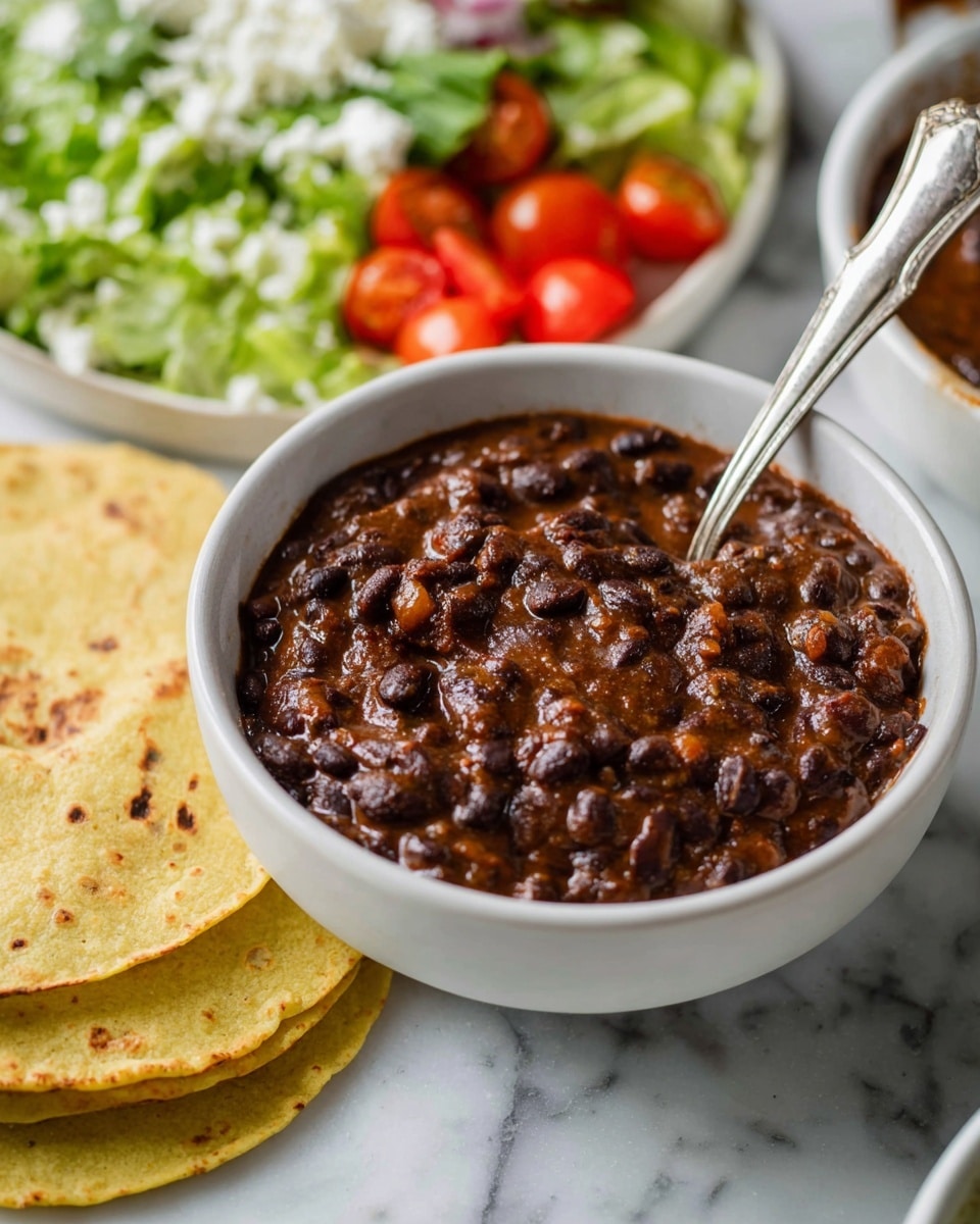 A white bowl filled with thick, dark brown-black beans mixed in a rich, slightly chunky sauce, with a shiny silver spoon standing in the middle. Around the bowl, there are two toasted, light yellow corn tortillas with small brown spots. In the blurred background, a white plate topped with layers of dark cooked beans, bright green lettuce, white cheese crumbles, and halved red cherry tomatoes adds color. The whole scene is set on a white marbled surface. Photo taken with an iphone --ar 4:5 --v 7