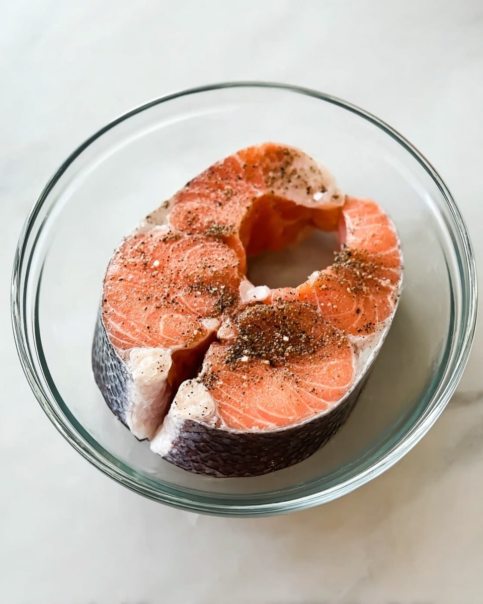 A clear glass bowl holds two raw salmon steaks inside, each with a thick layer of pink-orange flesh and a darker, rough-textured skin on one side. The salmon is seasoned with a light sprinkle of black pepper and some coarse grains of salt scattered evenly on top. The bowl sits on a white marbled surface, with soft lighting highlighting the fresh, raw texture of the fish. photo taken with an iphone --ar 4:5 --v 7