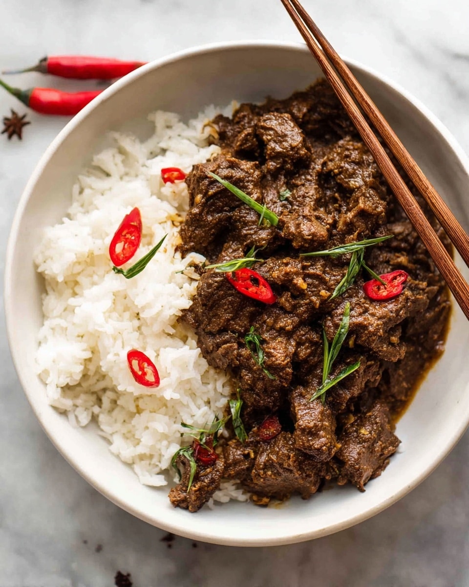 A white bowl filled with two main layers: on one side, there is a thick, dark brown meat stew with a coarse texture, topped with small slices of red chili and scattered thin green herb strips; on the other side, there is a fluffy white rice layer with visible grains, slightly mixed at the border with the stew. The bowl rests on a white marbled surface, and a pair of wooden chopsticks leans on the bowl from the top right corner. Photo taken with an iphone --ar 4:5 --v 7