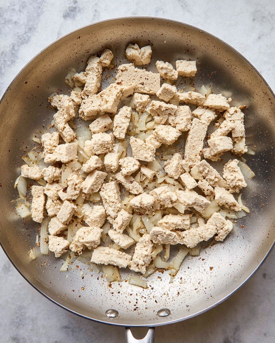 In a silver pan on a white marbled surface, there are small, uneven pieces of cooked light beige tofu with a slightly browned edge, mixed with small chunks of soft, translucent white onions. The tofu pieces vary in size and shape, scattered across the pan with some browned spots showing where it has cooked. The mixture spreads thinly and looks soft and lightly seasoned. Photo taken with an iphone --ar 4:5 --v 7