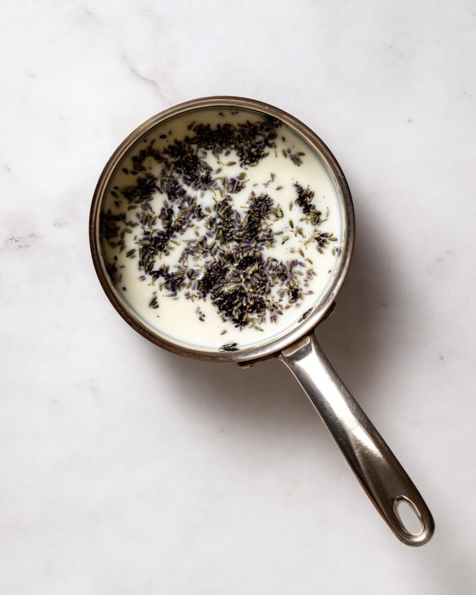 A small stainless steel pan with a long handle is placed on a white marbled surface. Inside the pan, there is a single layer of white creamy liquid topped with scattered dried lavender flowers, which are dark purple to black in color and unevenly spread across the surface. The pan reflects some light, showing a smooth metallic texture, and the white creamy liquid contrasts sharply with the dark lavender pieces. Photo taken with an iphone --ar 4:5 --v 7