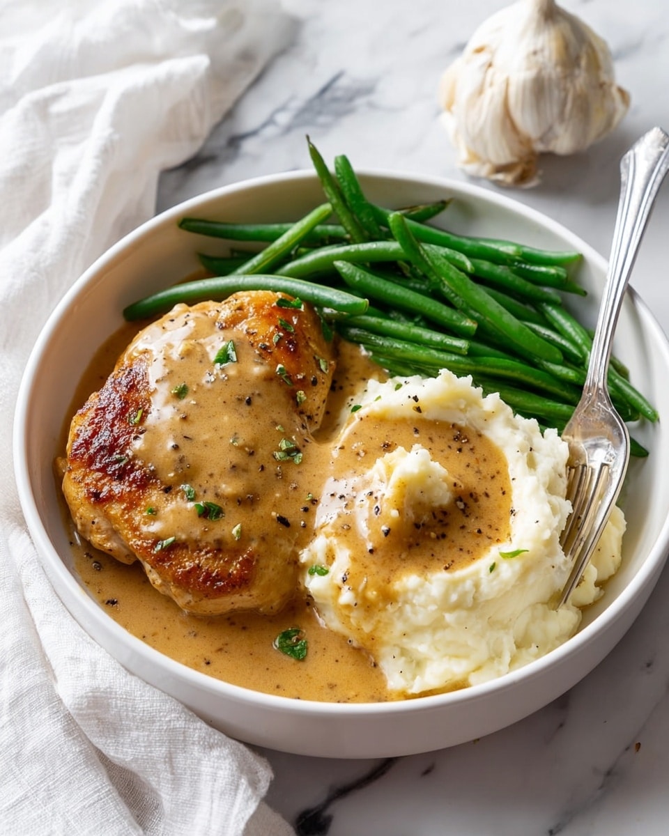 The dish shows a white bowl filled with three main parts: on the left, a golden-brown cooked chicken piece covered in light brown gravy with visible herbs; on the right, a mound of creamy white mashed potatoes topped with a pool of the same light brown gravy sprinkled with black pepper; above the mashed potatoes, a neat pile of vibrant, shiny green beans. A silver fork rests on the right side of the bowl, partially inside the mashed potatoes. The bowl is placed on a white marbled surface, with a garlic bulb and white cloth visible in the background. Photo taken with an iphone --ar 4:5 --v 7