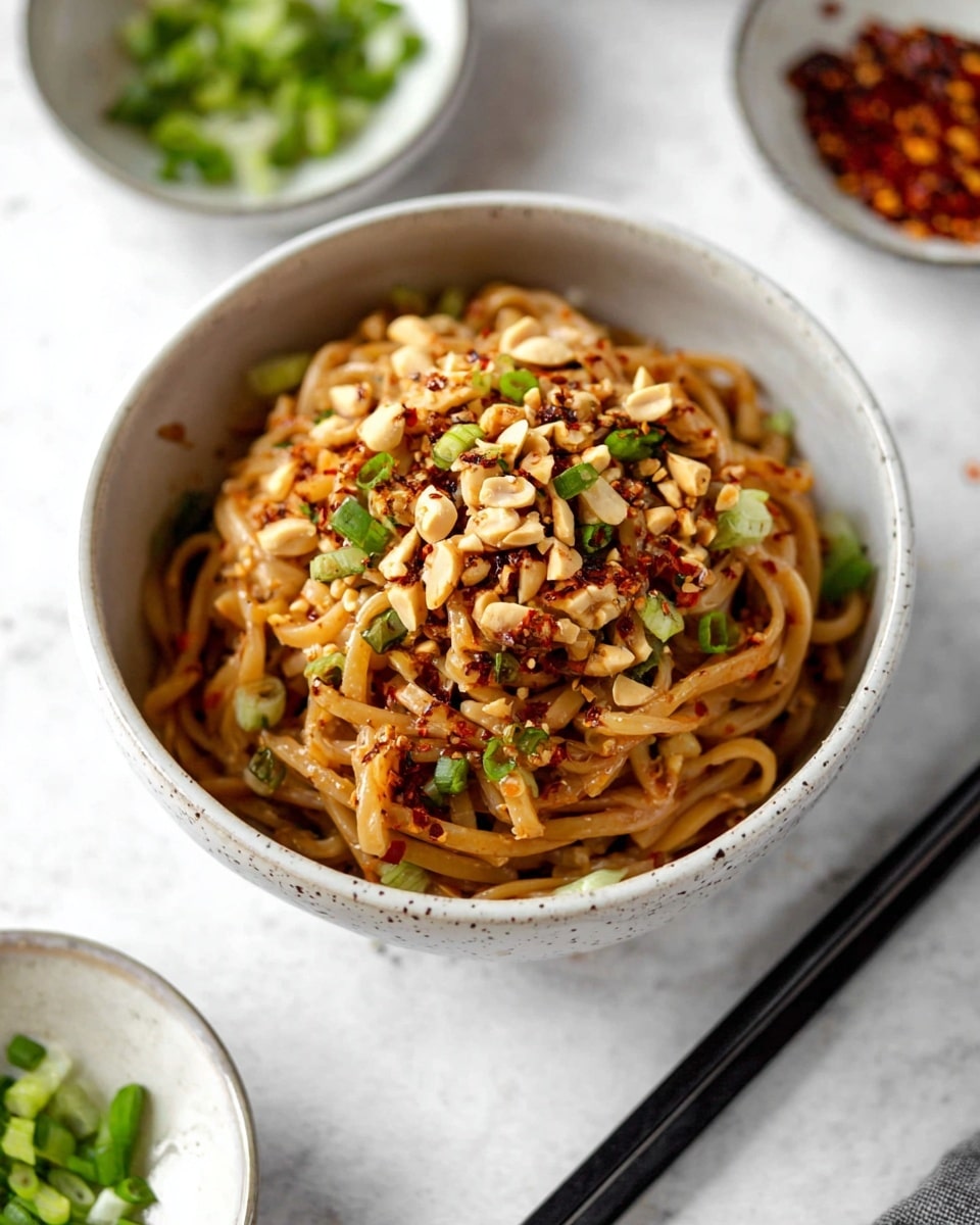 A bowl filled with thick brown noodles layered at the bottom, topped with light green sliced scallions scattered across, and chopped light tan peanuts sprinkled on top. The noodles have a slightly glossy texture with some red chili flakes distributed evenly, adding a touch of red color. The bowl is white with small gray speckles, sitting on a white marbled surface. Black chopsticks lie next to the bowl, and there are small white bowls in the background holding more sliced scallions and chili flakes. Photo taken with an iphone --ar 4:5 --v 7